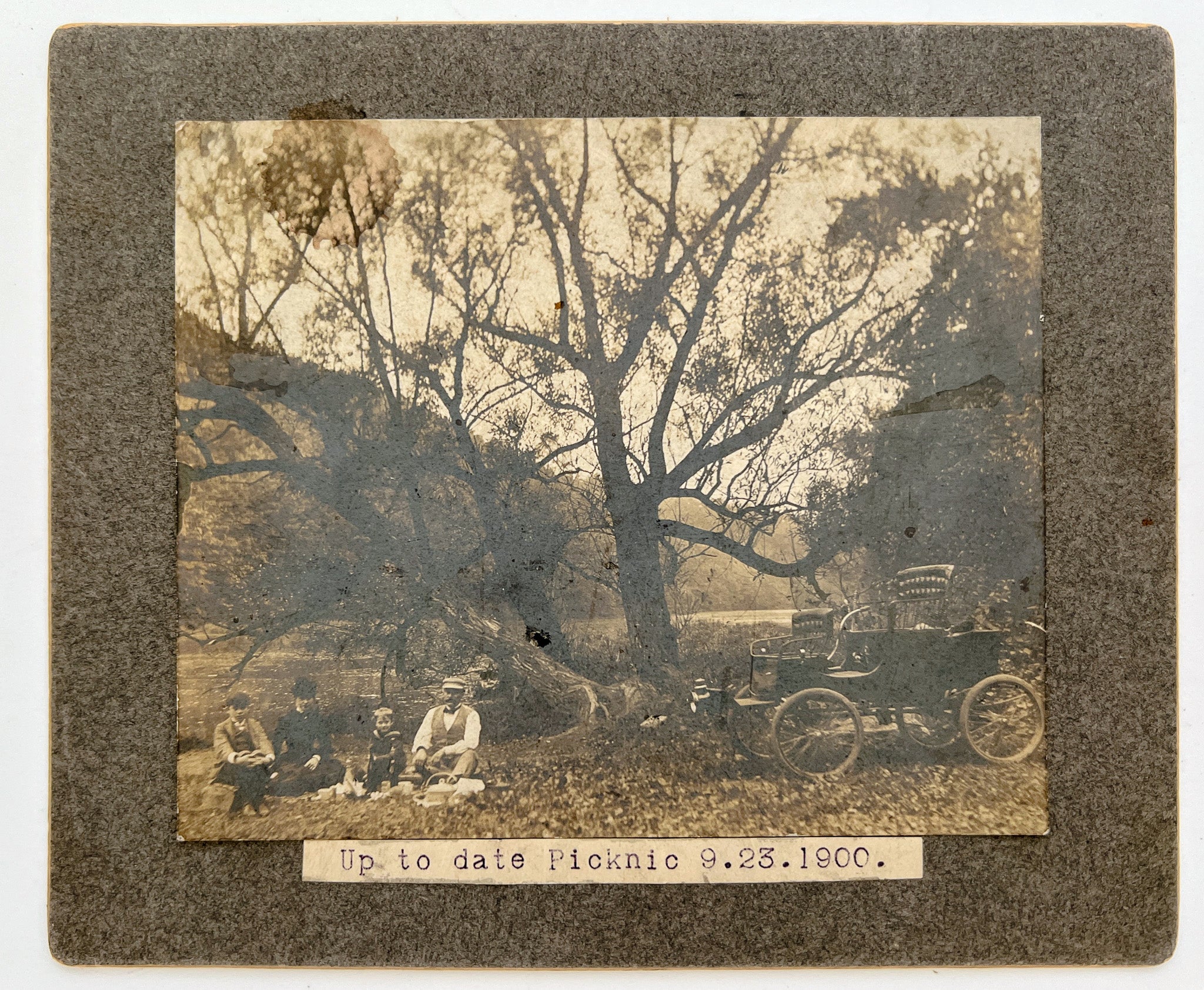 Mounted photo of a riverside family picnic with early automobile, 1900