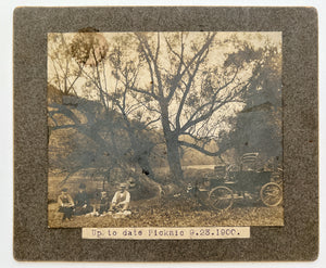 Mounted photo of a riverside family picnic with early automobile, 1900
